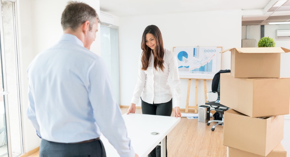 An older white male dressed in business clothes moving an ergonomic white office desk with a young white female colleague wearing office clothes in a well lit large office space