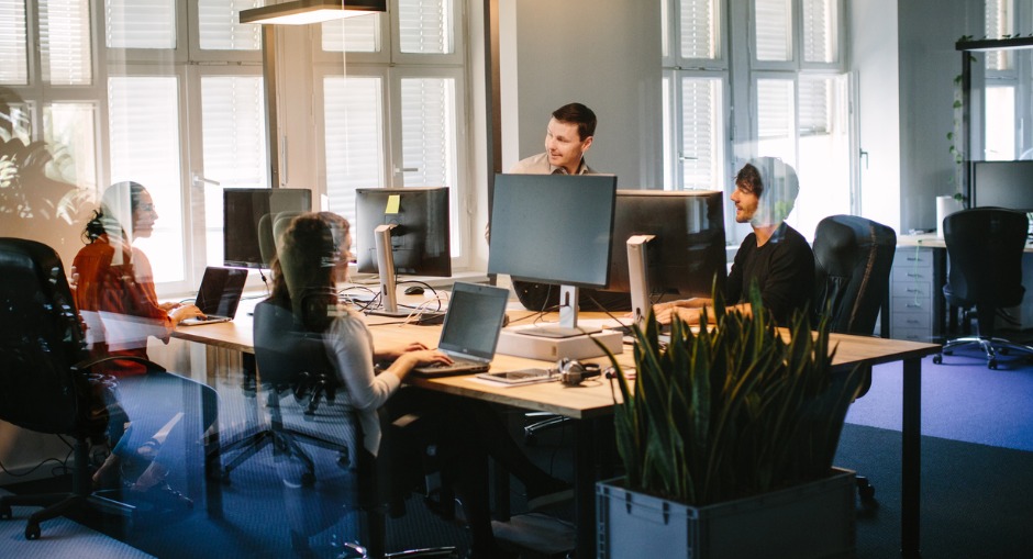 Four colleagues sitting in an office at a communal desk with computers in front of them and overhead office lighting.